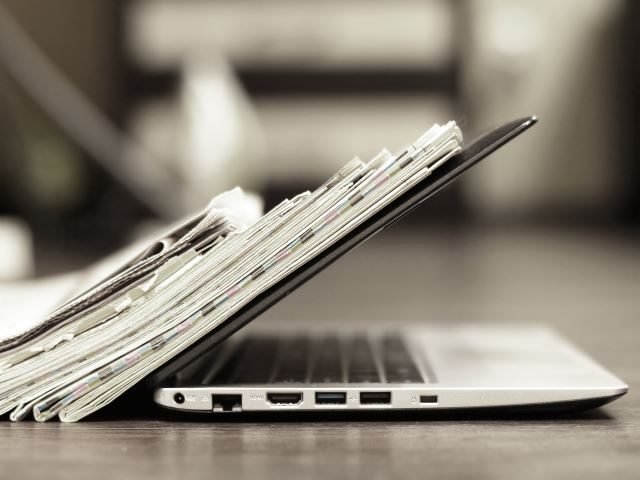Open laptop with a stack of newspapers leaned up against it - Newsbyte concept